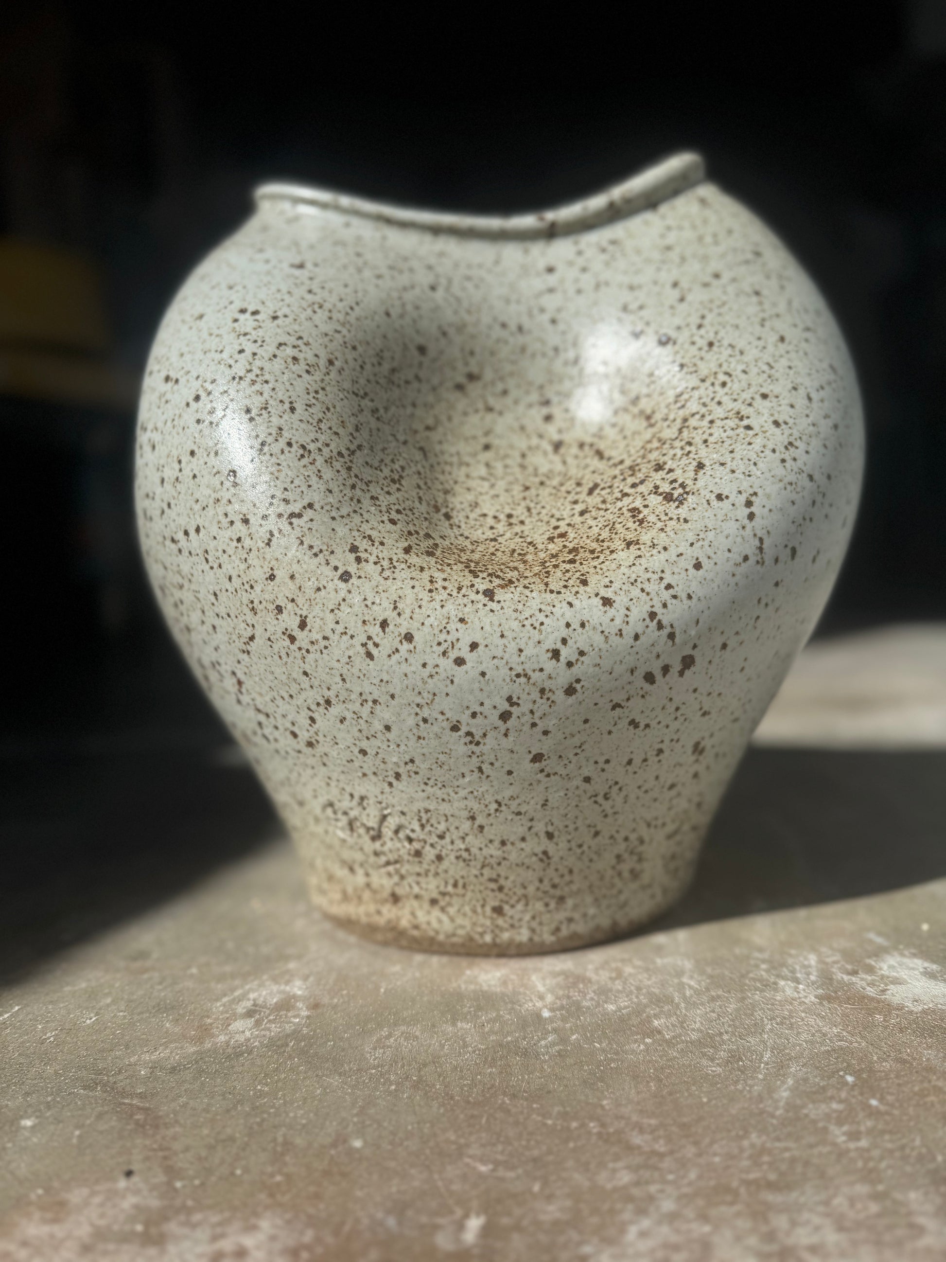 A hugged ceramic vase with a rounded, bulbous shape and a small, irregularly shaped rim, featuring a speckled beige and brown glaze.