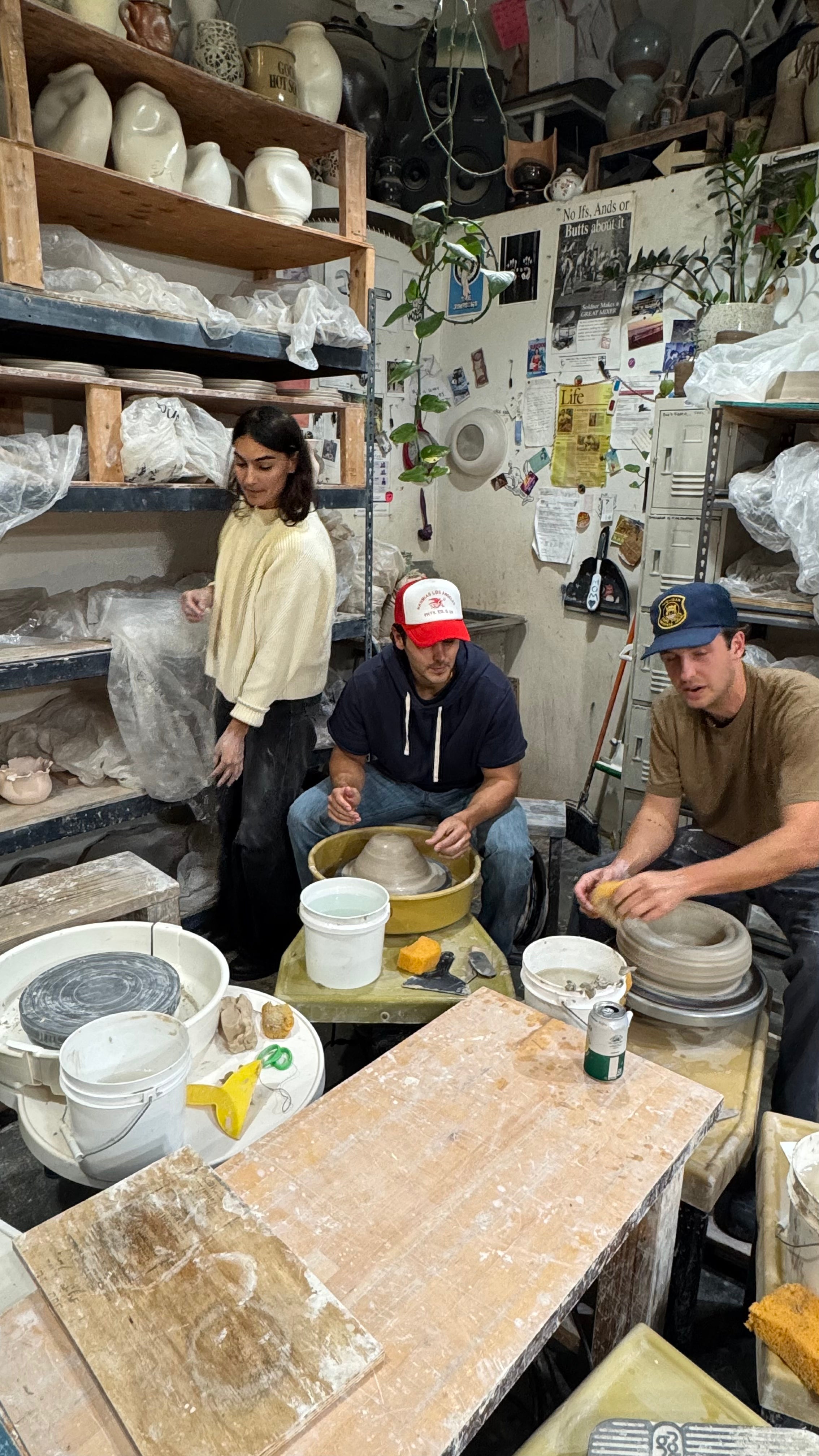 Three people working at pottery wheels in a cluttered but creative studio filled with clay, shelves, and tools.
