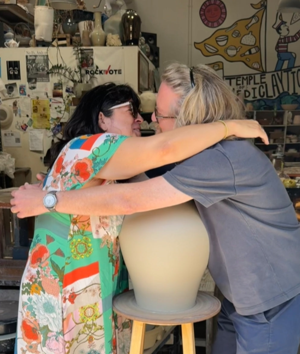 Two people embracing in a pottery studio, with a large cream-colored ceramic vase on a wooden stool in the foreground. The background shows shelves with pottery and colorful wall art.